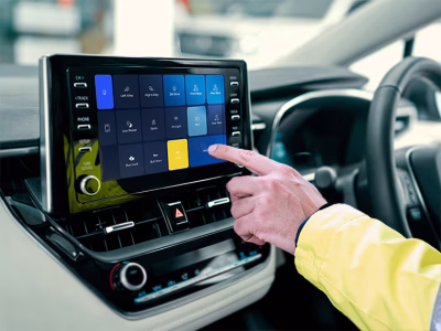 UniScreen OEM Touchscreen integration shown on Toyota Corolla infotainment screen with dashboard and steering wheel. Hand pressing screen with hi-vis cuff.