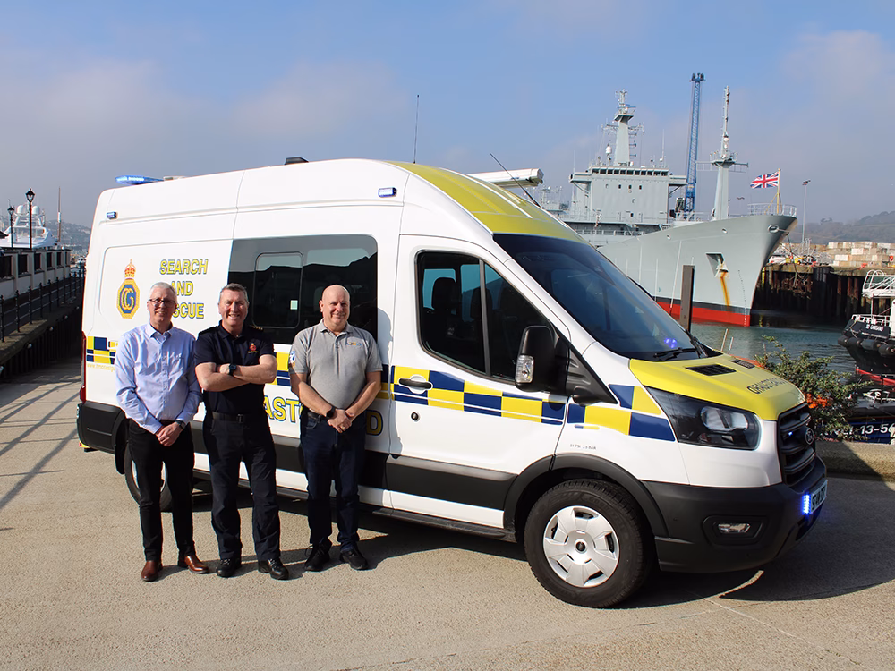 HM Coastguard Ford Transit OSV with lit blue lights parked at an angle in a harbour on a bright day with a grey Naval Vessel in the background. A Standby and EVO spokesperson stand either side of a Coastguard representative.