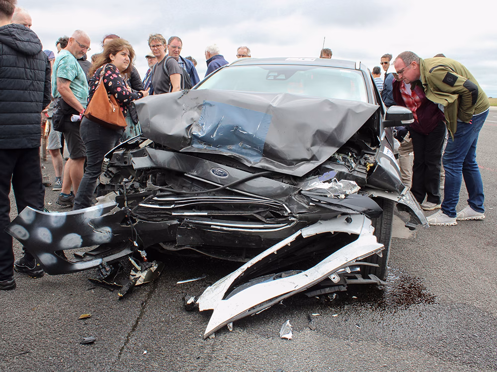 Aftermath of a staged crash with heavily damaged black vehicle and remnants of a lighter vehicle's bumper taken on an airfield runway and surrounded by onlookers at ITAI Crash Day 2026