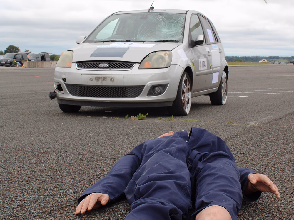 Silver ford with broken windscreen wnd crash test dummy sprwaled on floor in foreground at ITAI Crash Day 2026
