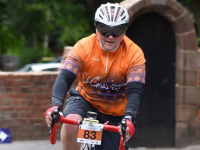 Standby UK's Lee Beaumont cycling on a racing style bike, wearing an orange St Giles t-shirt in front of a stone archway / door and wall on a UK street.