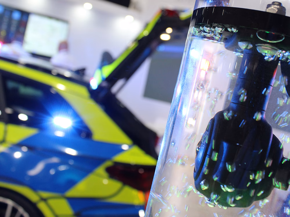 Close up angle shot of an Oculux camera in a lit up bubble tube, a South Wales Police Cupra Leon Estate RPU converted by Elite Solutions is out of focus on the Standby stand at NAPFM 2026.
