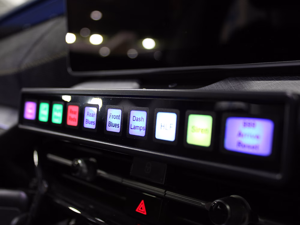 Closely cropped MCS-F5D Slimline Switch Unit shown mounted on a vehicle dash which is out of focus, the three centremost buttons are in focus with their various functions shown against colurfully lit backgrounds, the rest are out of focus.