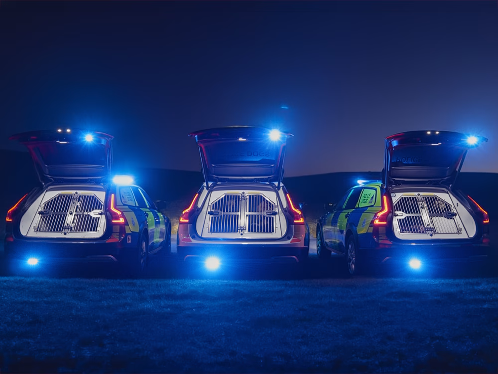 Three Volvo police dog units parked side-by-side at night with their rear hatches open, revealing metal dog crates, with their blue emergency lights illuminated in synchronisation.