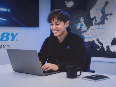 A smiling worker sits at a white desk working at a laptop in a modern office space in front of a map wall graphic. A picture of a lightbar is cropped in the corner and part of the Standby logo underneath it on the wall. They're wearing a black fleece with a blue Standby S, smiling and looking at the screen as they type.
