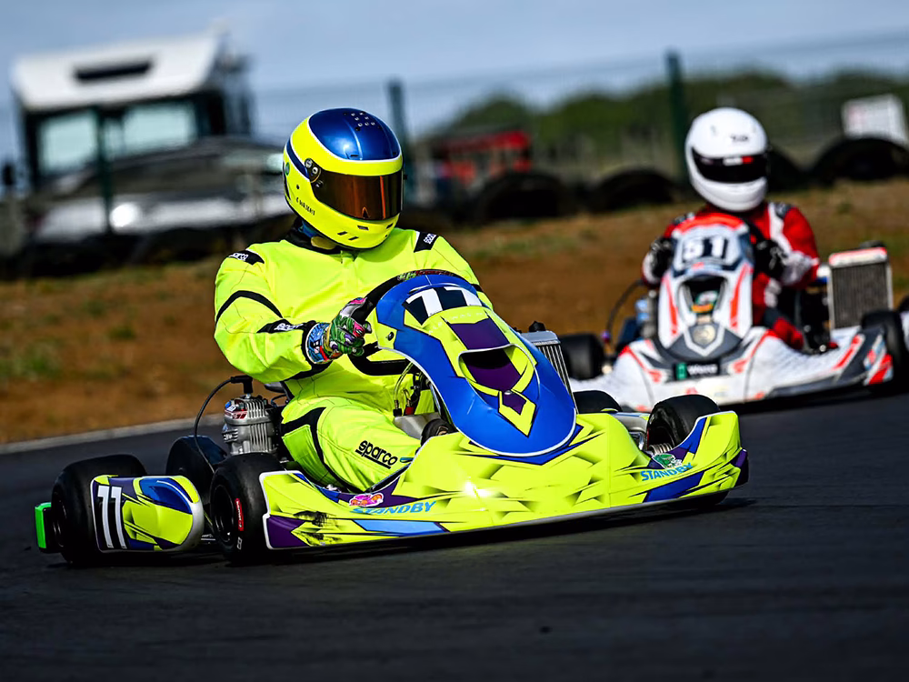 Go kart racer (nuber 77) in fluorescent yellow overalls and matching kart with dark blue accents driving on a tarmac track with grass and tyre wall in front of a chain link fence in the background, on a bright clear day. Racer 51 is in red and white slightly behind 77, cornering round a bend.