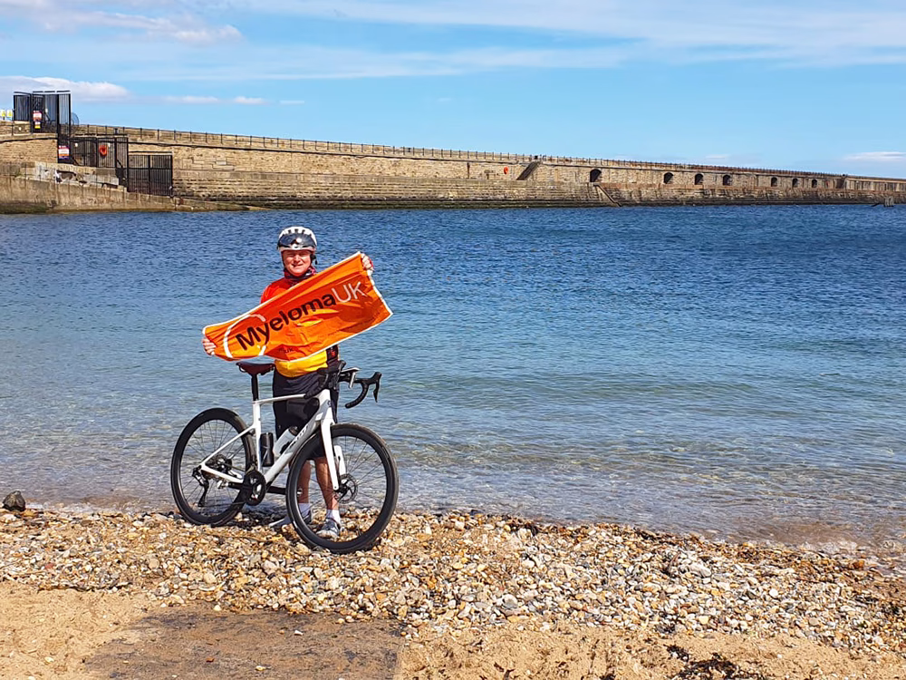 A cyclist in a helmet stands on a pebbly beach, holding an orange Myeloma UK banner next to a white bicycle, with blue water and a long stone pier in the background.