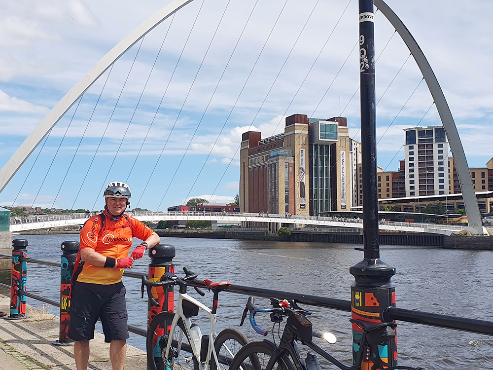 A cyclist in an orange jersey stands by two bikes on a riverside path, with a large arch bridge and modern buildings in the background under a partly cloudy sky.