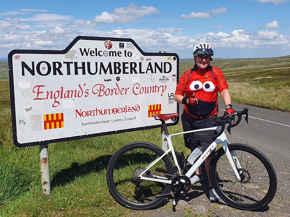 A cyclist wearing a red Elmo jersey stands beside a bike and a Welcome to Northumberland sign on a sunny rural road, with green hills and blue sky in the background.