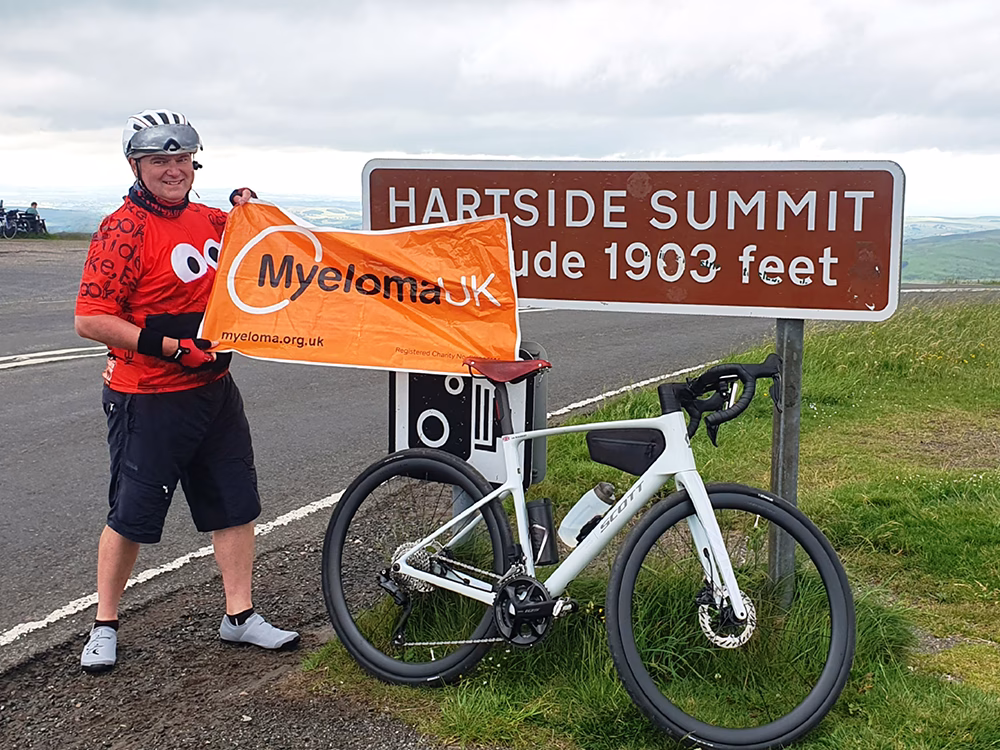 A smiling cyclist in a red shirt holds a Myeloma UK banner next to a bike and the Hartside Summit sign, which reads altitude 1903 feet, with hills and cloudy sky in the background.