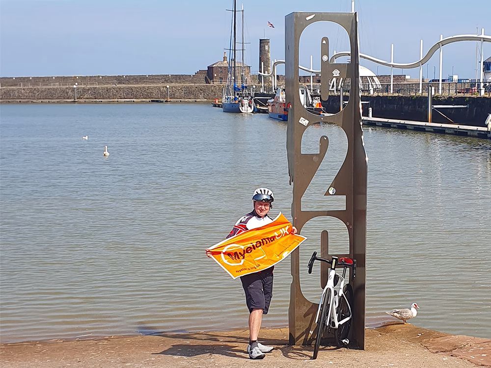 A cyclist stands by a metal C2C sculpture near a harbor, holding a myeloma UK flag. A bike is parked beside them, with boats, water, and a water bird in the background under a clear sky.