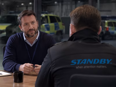 Modern corporate meeting room interior, a wide window in the back reveals modern British Police Car fleet in a garage out of focus and quite dark, two people sit at a table in the meeting room, the one closest to the foreground has their back to the camera and wears a black softshell jacket with the Standby 'when attention matters' logo, the other is facing them, wearing smart casual attire
