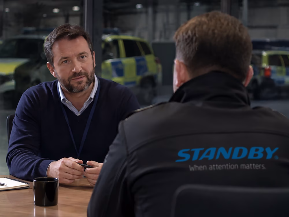 Modern corporate meeting room interior, a wide window in the back reveals modern British Police Car fleet in a garage out of focus and quite dark, two people sit at a table in the meeting room, the one closest to the foreground has their back to the camera and wears a black softshell jacket with the Standby 'when attention matters' logo, the other is facing them, wearing smart casual attire